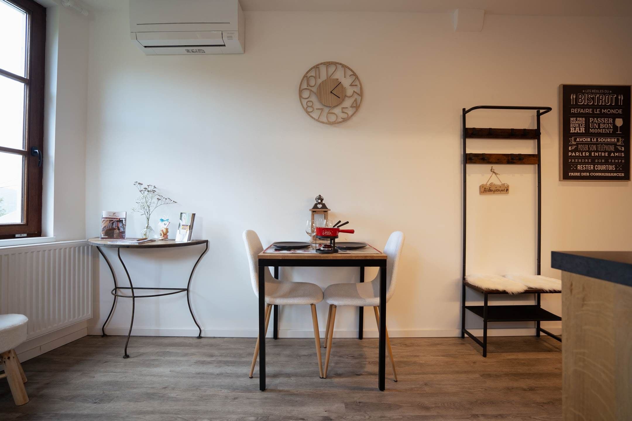Ein Zimmer mit Esstisch, zwei Stühlen, einer Wanduhr, einer Heizung, einem Fenster und einem an der Wand befestigten Regal.