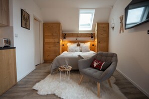 A bedroom with a bed, a chair, a small table, a fluffy rug, and a TV on the wall. - Le Petit Chalet: 2-person vacation rental with spa access (AUBURE)