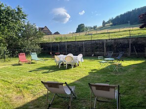 A grassy outdoor area with a stone wall, a table with chairs, and a few folding chairs. - L'Orée des Sapins: 2-person vacation rental with spa access (AUBURE)