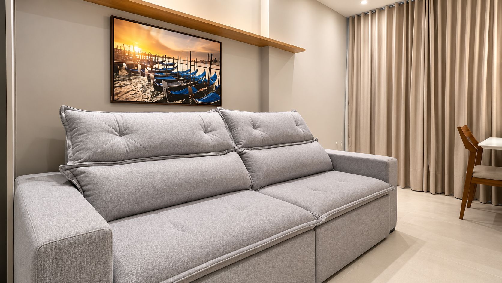 Un canapé gris avec un coussin, une chaise en bois et un tableau représentant des bateaux accroché au mur.