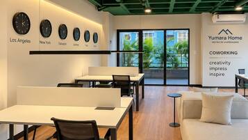 Espaço de trabalho moderno com mesas, cadeiras e um sofá. Há relógios indicando diferentes cidades e uma parede com a marca 'Yumara Home'.