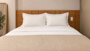 Cama com cabeceira de madeira, roupa de cama branca e manta bege texturizada.