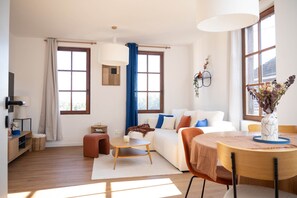 Un salon moderne avec un canapé, une table basse et une table à manger. Il y a deux fenêtres avec des rideaux, un luminaire fixé au mur et une petite table d’appoint.