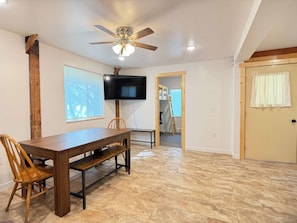 Un comedor con mesa y sillas de madera, ventilador de techo, televisor y puerta que da a otra habitación.