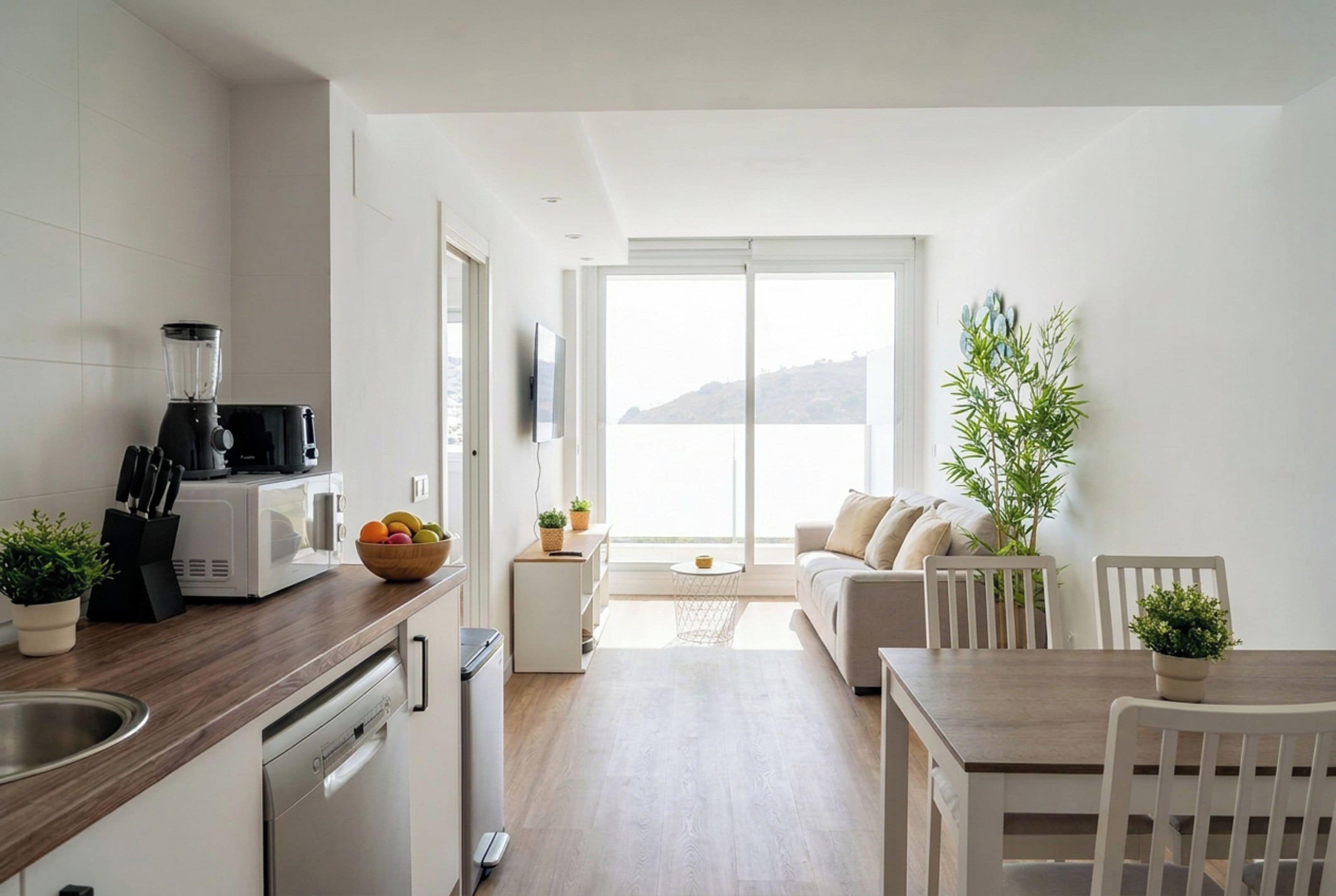 A modern kitchen with a blender, microwave, and dishwasher, leading to a balcony with a view.