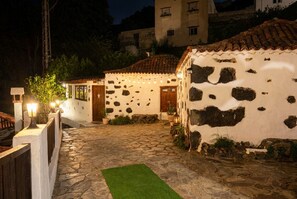 Une cour dallée de pierres, avec des bâtiments à l’aspect rustique, une clôture en bois et des guirlandes lumineuses.