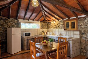 A rustic kitchen with a stone wall, wooden ceiling beams, a tiled backsplash, a refrigerator, a microwave, a sink, and a dining table with chairs. - Las Ninfas Natural Escape 3 (Icod de los Vinos)