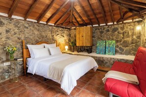 A bedroom with a stone wall, a wooden ceiling, a bed with white linens, a red chair, and a small table with a lamp. - Las Ninfas Natural Escape 3 (Icod de los Vinos)