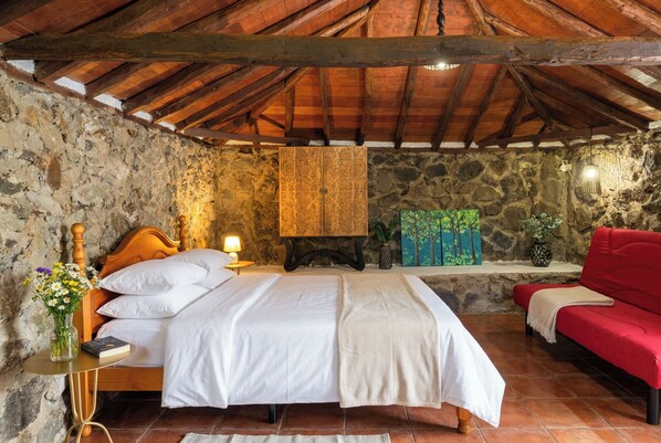 Une chambre avec un mur en pierre, un lit en bois, un canapé rouge et un tableau accroché au mur.