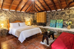 Une chambre avec un mur en pierre, un plafond en bois, un lit avec du linge de lit blanc, une petite table avec une plante et une chaise rouge.