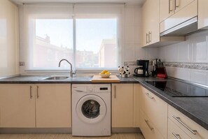 A compact kitchen with a washing machine, a sink, and a window with a view of buildings. - Corralejo 3BR House with Pool (Corralejo)