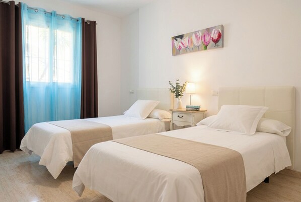 A hotel room with two beds, a window with curtains, a painting on the wall, and a bedside table with a lamp. - Corralejo 3BR House with Pool (Corralejo)