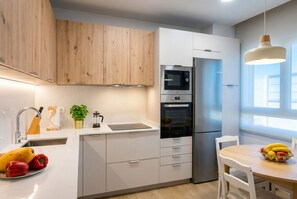 Une cuisine moderne avec des armoires blanches, un réfrigérateur en acier inoxydable, un four et un four à micro-ondes. On y trouve un évier avec un robinet, une plante en pot et un panier de fruits sur le comptoir.