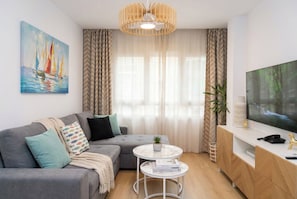 Ein modernes Wohnzimmer mit einem grauen Sofa, einem kleinen runden Tisch, einem Fernseher auf einem Holzschrank und einem Segelboot-Bild an der Wand.