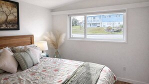 A bedroom with a bed, a nightstand, a lamp, a vase with dried flowers, a ceiling fan, and a window with a view of a building. - Seaview Suite · sunrise deck, BBQ, firepit & 3-bed (Campbell River)