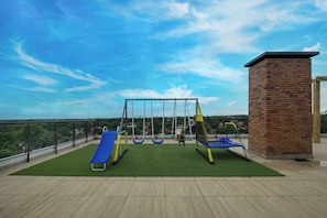 A rooftop playground with swings and a slide, surrounded by a brick wall and a metal railing. - Cozy apt relaxing view - Great Guest Service 604 (Fernando de la Mora)