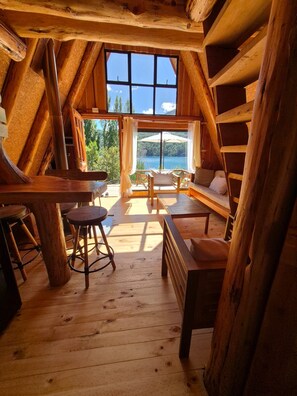 A wooden cabin with a large window overlooking a lake, furnished with a sofa, chairs, and a table.