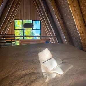 An attic room with a wooden sloped ceiling, a large window with a view of trees, and an open book on a bed.