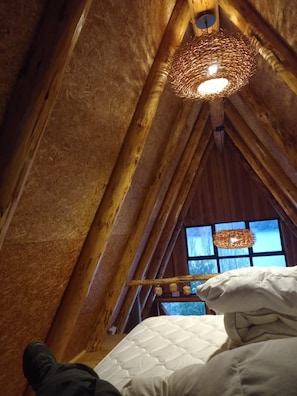Una habitación con techos altos y un techo de madera inclinado, una cama con ropa blanca y una lámpara de fibra.