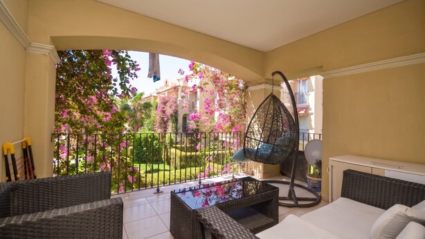Ein Balkon mit Korbmöbeln, einem Hängesessel und Blick auf einen Garten mit rosa Blüten.