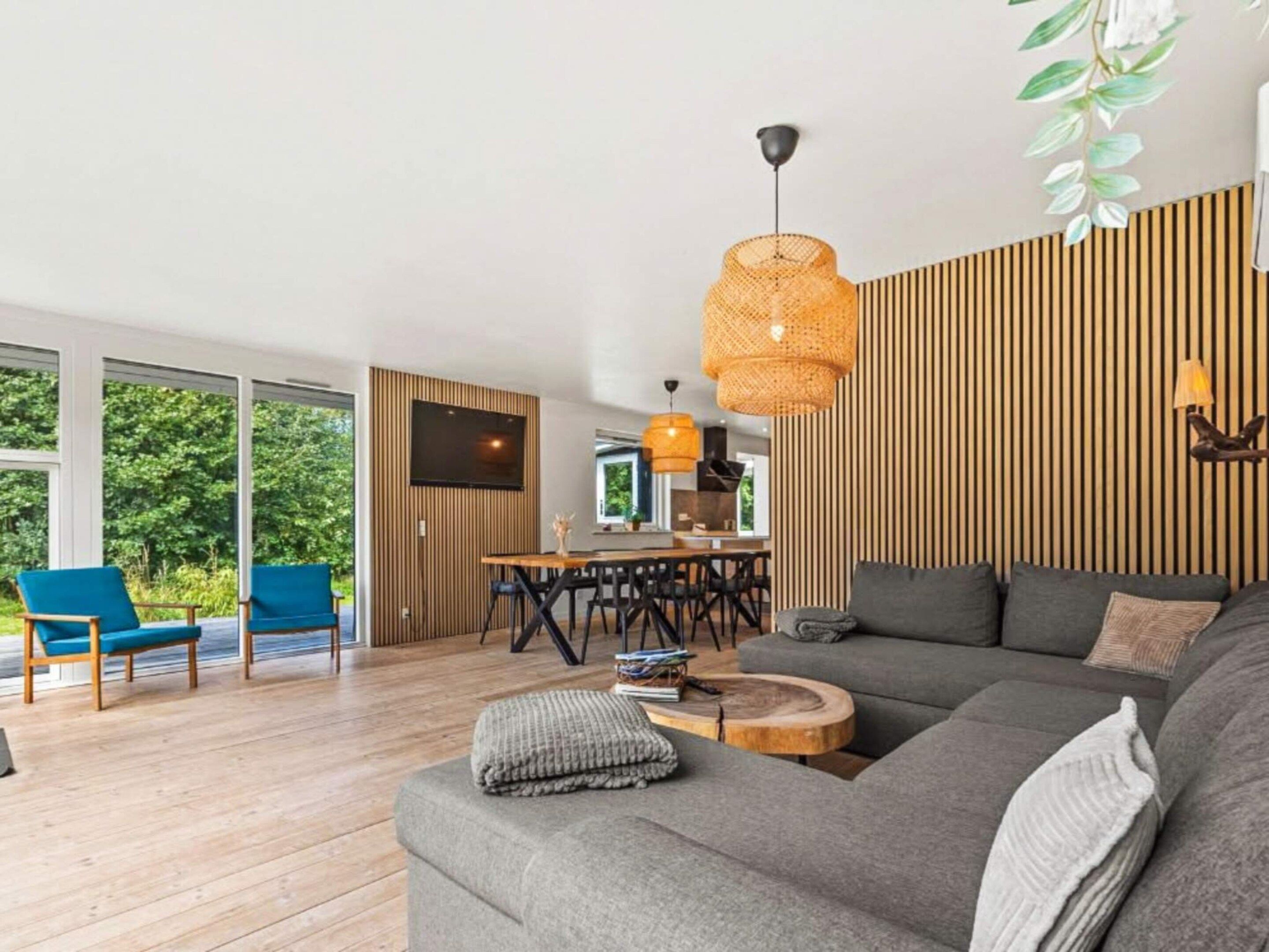 A modern living room with a grey sofa, blue chairs, a wooden dining table, and a large window with a view of greenery.