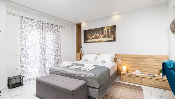 A modern bedroom with a large bed, a bedside table, a nightstand, a bench, and a painting on the wall. - Premium Suite with veranda and panoramic sea view, Notus Hotel Chania (Chania)