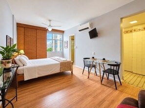 A hotel room with a bed, a desk with chairs, a ceiling fan, an air conditioning unit, and a TV on the wall.