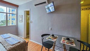 Habitación de hotel con cama, mesa con sillas, equipo de aire acondicionado, televisión y ventana con vista a edificios.