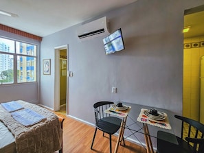 Habitación de hotel con cama, mesa con sillas, equipo de aire acondicionado, televisión y ventana con vista a edificios.