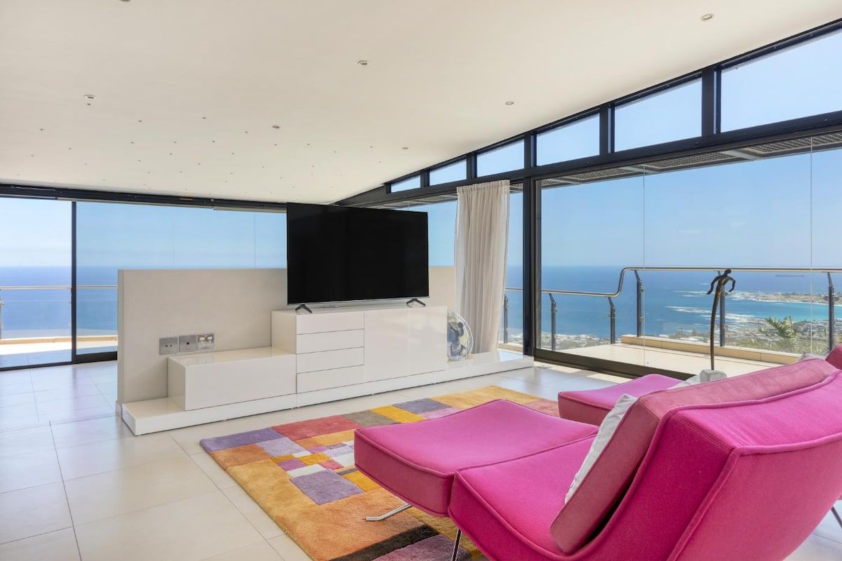 Un salon moderne avec un grand téléviseur, un balcon offrant une vue sur la mer et des fauteuils de détente roses.