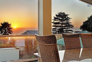 A balcony with wicker chairs and a table, overlooking a sunset view with trees and buildings. - Walking distance to Camps Bay Beach, Views - Merridew (Cape Town)