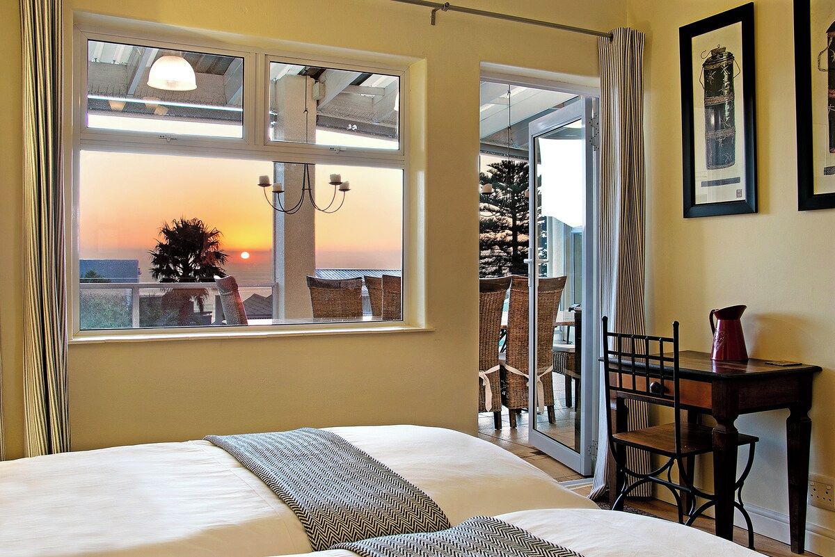 A hotel room with a bed, a window with a view of the sunset, a dining area with chairs, and a table with a pitcher.