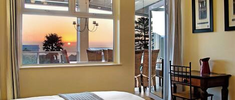 A hotel room with a bed, a window with a view of the sunset, a dining area with chairs, and a table with a pitcher.