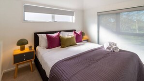Een slaapkamer met een bed, kussens, een nachtkastje, een lamp en een raam.