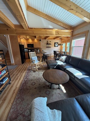 A spacious living area with a wooden ceiling, a black leather sofa, a patterned rug, and a dining area with a table and chairs.
