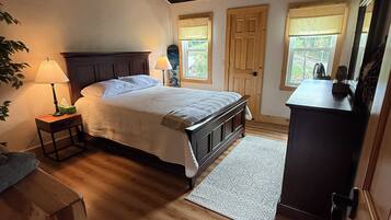 A bedroom with a wooden floor, a bed with a headboard, a nightstand, a lamp, a dresser, and a window with a view.