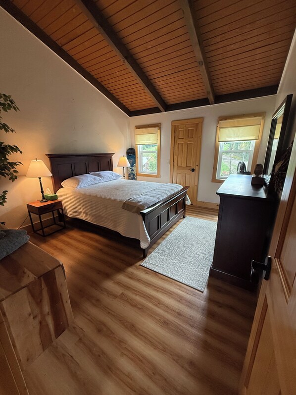 A bedroom with a wooden floor, a bed with a headboard, a nightstand, a lamp, a dresser, and a window with a view.
