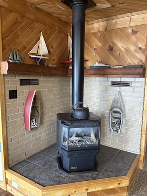 A wood-burning stove in a corner of a room with a brick wall, a wooden ceiling, and a small shelf with model ships. - Sandy beach, hot tub, amazing sunsets, spacious lake home, kayaks and more. (Wautoma)