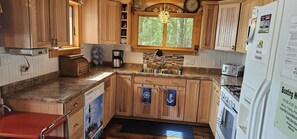 A compact kitchen with wooden cabinets, a built-in sink, a stove, and a window with a view of trees. - Sandy beach, hot tub, amazing sunsets, spacious lake home, kayaks and more. (Wautoma)
