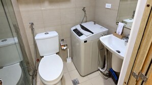 A compact bathroom with a toilet, a washing machine, a sink, and a glass shower enclosure. - Shore 1 Residences by LUXE (Pasay City)