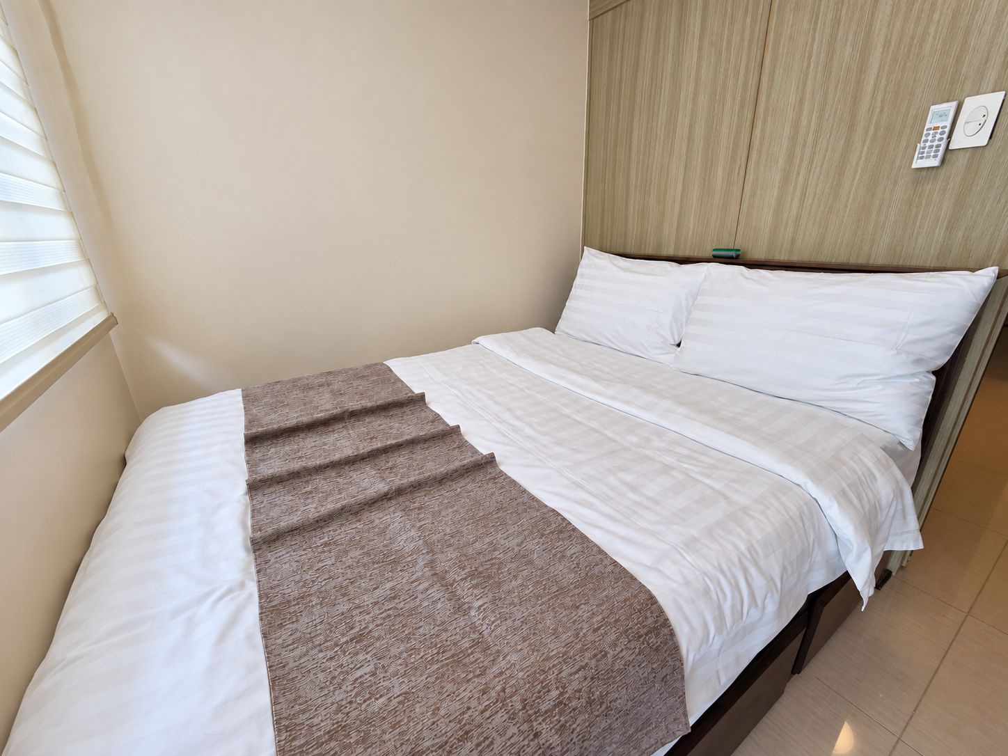 Una cama individual con ropa de cama blanca y una manta marrón, un cabecero de madera y un panel de control en la pared.