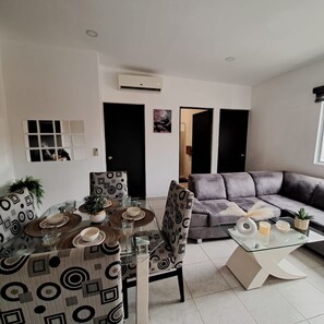 Uma sala de estar moderna com uma mesa de centro de vidro, poltronas estampadas, um sofá cinza e uma área de jantar com mesa e cadeiras de vidro.
