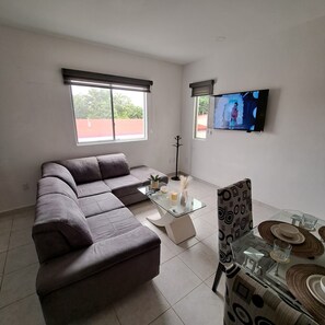 Uma sala de estar moderna com um sofá seccional, uma mesa de centro de vidro, uma mesa de jantar posta com pratos e copos, e uma TV de tela plana fixada na parede.