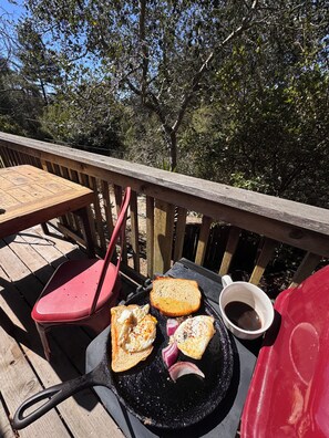 Een houten terras met een tafel en stoelen, een zwarte pan met toast en uien, en een mok.