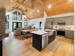 A modern kitchen with a large island, stainless steel appliances, and a stone accent wall. - Blue Ridge Serenity - Creekside | Hot Tub | Firepit | Wildlife Galore! (Morganton)