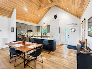 A modern kitchen with a central island, wooden ceiling, and a dining area with a table and chairs. - Blue Ridge Serenity - Creekside | Hot Tub | Firepit | Wildlife Galore! (Morganton)