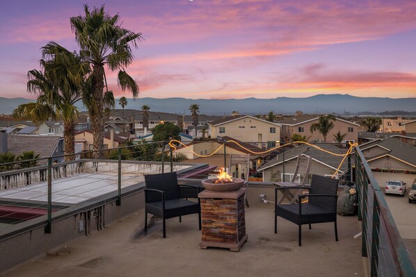 A rooftop patio with a fire pit, outdoor seating, and a view of a residential area with mountains in the background.