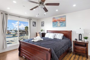 A bedroom with a wooden bed, a ceiling fan, a nightstand with a lamp, and a painting of a sunset over the ocean.