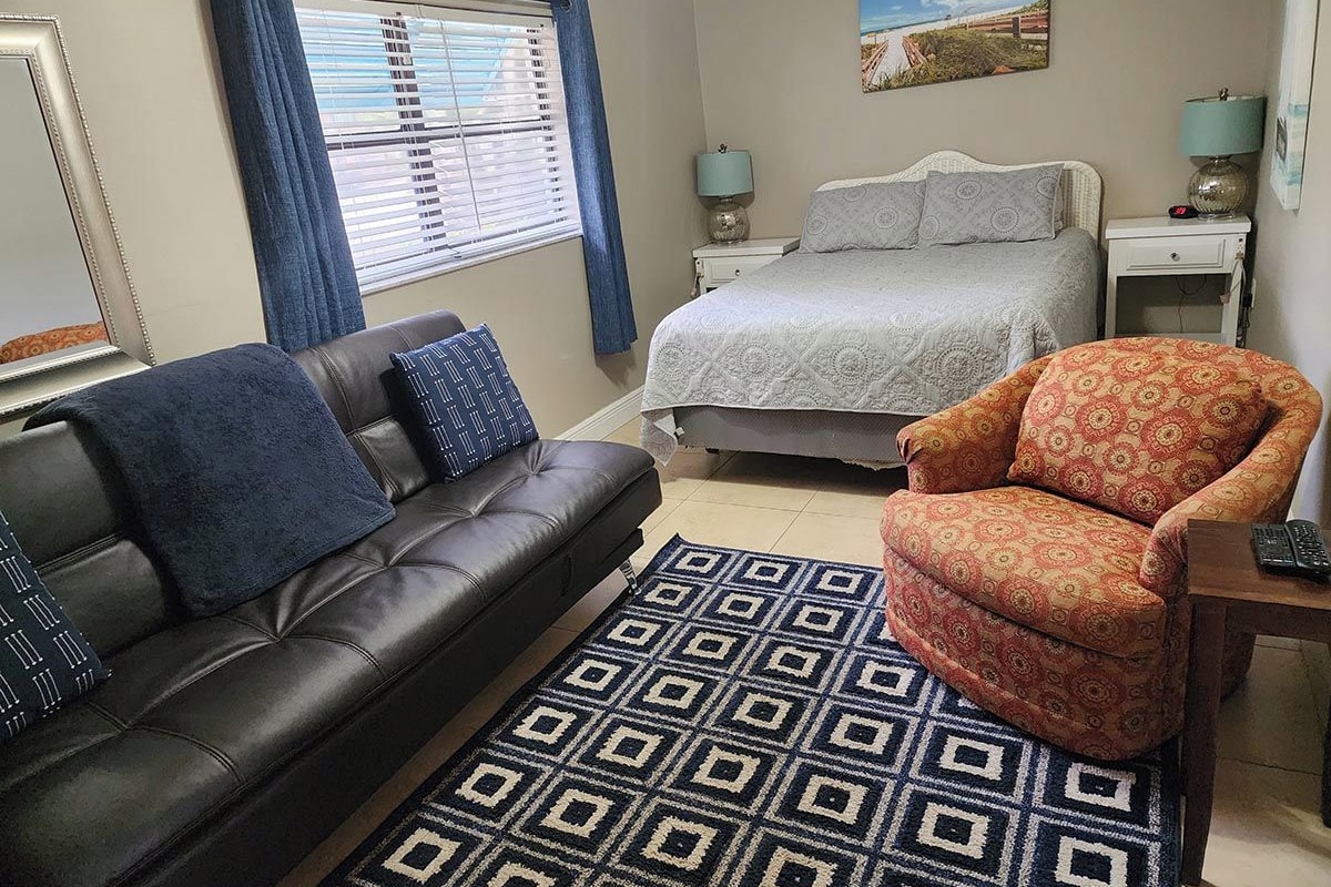 A bedroom with a bed, a sofa, a chair, a window with curtains, and a painting on the wall.