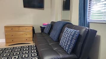 A room with a black leather sofa, a wooden dresser, a patterned rug, a television on the wall, and a window with curtains.